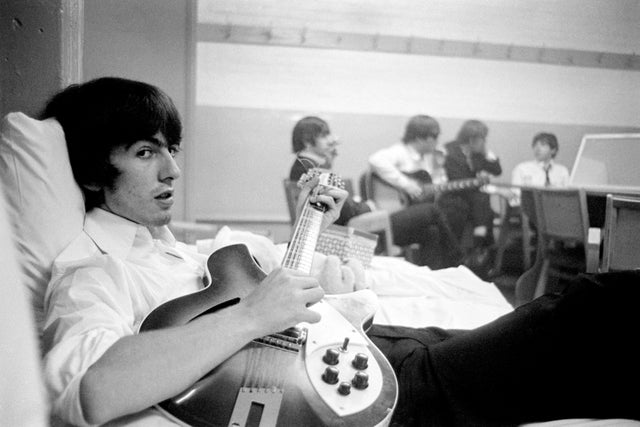 George Harrison deitado com sua guitarra, enquanto os outros Beatles conversam ao fundo, 1964.