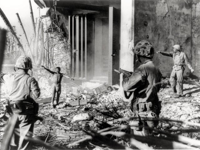 Um soldado japonês se rendendo a três fuzileiros navais dos Estados Unidos nas Ilhas Marshall, janeiro de 1944. Um momento tenso em que o prisioneiro deve primeiro se despir para mostrar que não deseja realizar um ataque suicida.