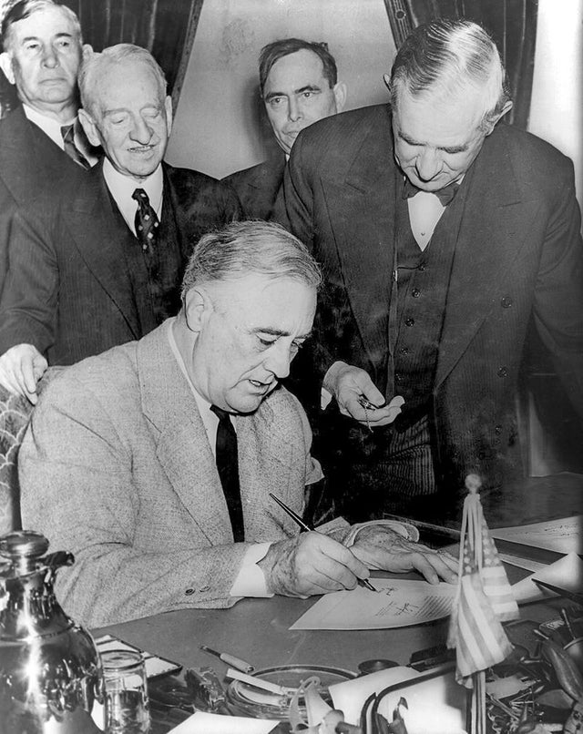 Presidente Franklin D. Roosevelt assinando a declaração de guerra contra a Alemanha, marcando a entrada dos Estados Unidos na Segunda Guerra Mundial. O senador Tom Connally é visto à esquerda de Roosevelt segurando um relógio para marcar a hora exata da assinatura, 1941.