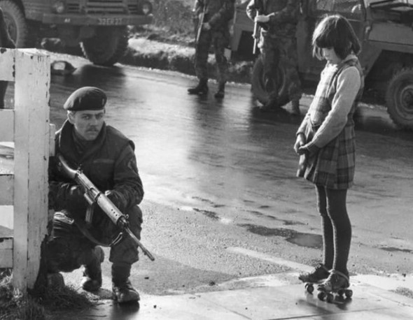 Um soldado britânico de guarda em Belfast, com uma garotinha curiosa de patins observando-o, 1971.