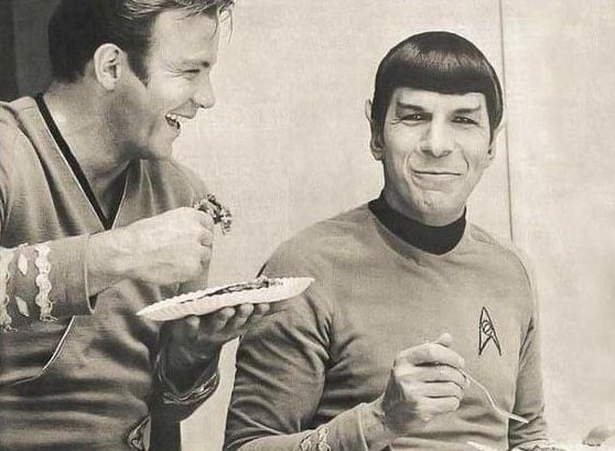 William Shatner comemorando seu aniversário dividindo um pedaço de bolo com um sorridente Leonard Nimoy.